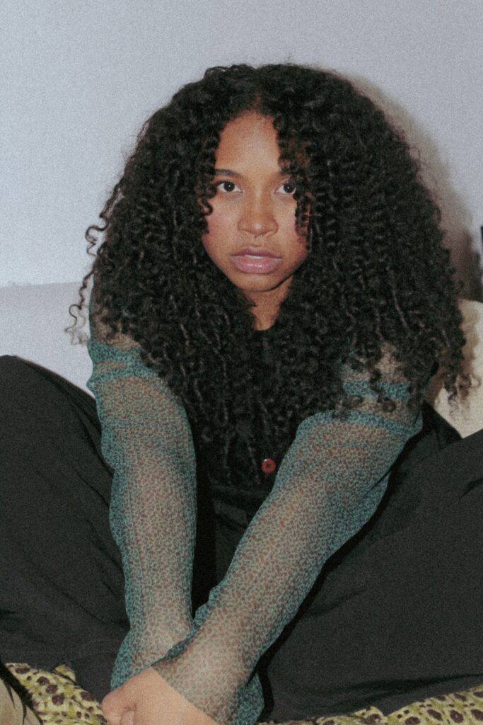 A young woman with long, black curls sits cross-legged on a sofa and looks directly into the camera.