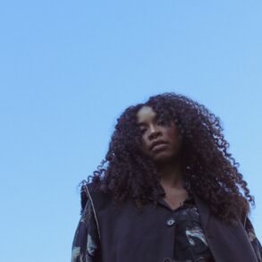 Eine junge Frau mit dichten, schwarzen Locken blickt von oben in die Kamera hinunter, über ihr blauer Himmel.