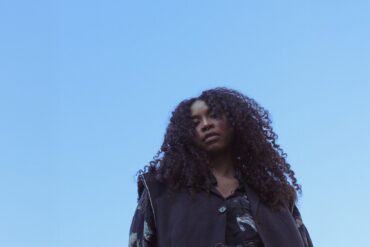 A young woman with thick, black curls looks down into the camera from above, blue sky above her.