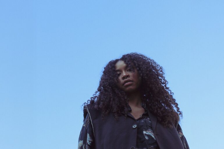 A young woman with thick, black curls looks down into the camera from above, blue sky above her.
