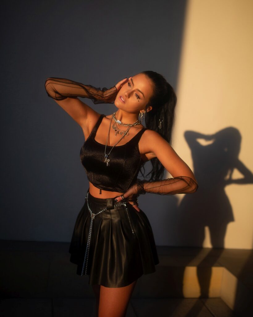 Eine junge Frau mit dunklem Haar im Zopf, schulter- und bauchfreiem schwarzen Top und schwarzem Minirock steht vor einer weißen Wand und wird vom Sonnenuntergang angestrahlt.
