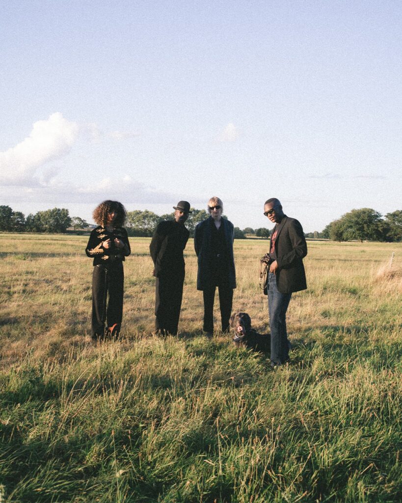 A young woman and three young men are standing next to each other on a lawn, dressed smartly, and looking at the camera. A dog is also lying next to them in the grass.