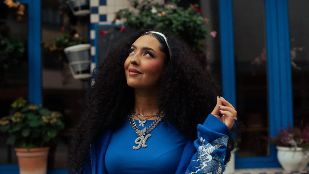 Eine junge Frau mit dichtem, schwarzen Haar, das von einem Haarreif zurückgehalten wird, steht vor einem Gebäude und blickt nach links oben. Sie trägt ein blaues Oberteil und eine Metallkette, an der ein Anhänger "K" hängt.