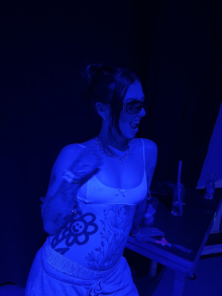 A young woman with dark hair and sunglasses stands in a dark room, illuminated only by blue light. She is wearing a bra and sweatpants, and her stomach and arms are covered in numerous tattoos.