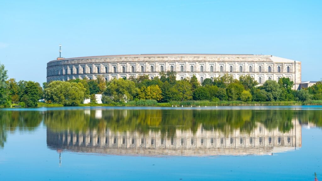 Hinter einem See und einer Reihe Bäume steht ein riesiges, weißes, zweigeschossiges Gebäude, das sich in der stillliegenden Wasseroberfläche spiegelt.