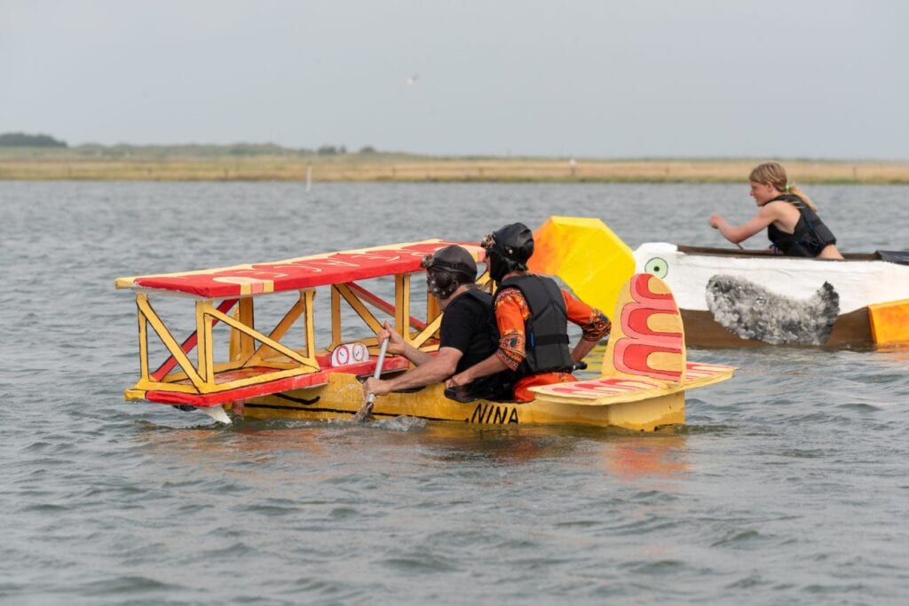 Zwei Personen sitzen in einem aus Papier nachgebauten Doppeldeckerflugzeug und paddeln über eine Meeresbucht. Hinter ihnen sitzt eine junge Person in einem Papierboot, das eine Möwe darstellt.