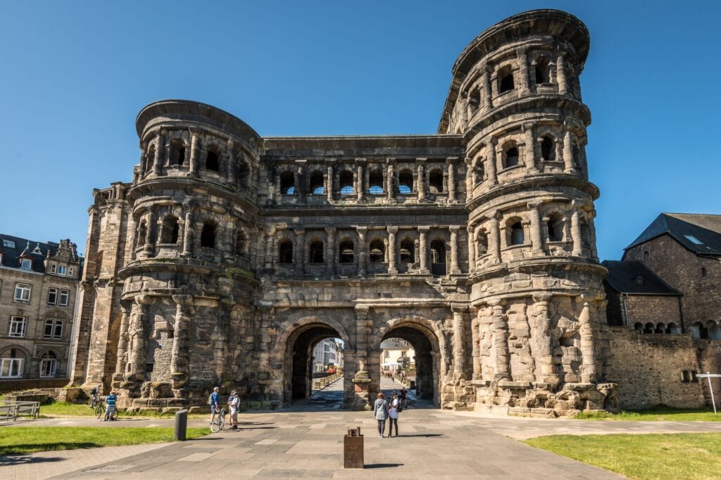 Die Porta Nigra, vom Norden aus gesehen. Auf der Straße davor sind einige Menschen unterwegs, der Himmel darüber ist blau.
