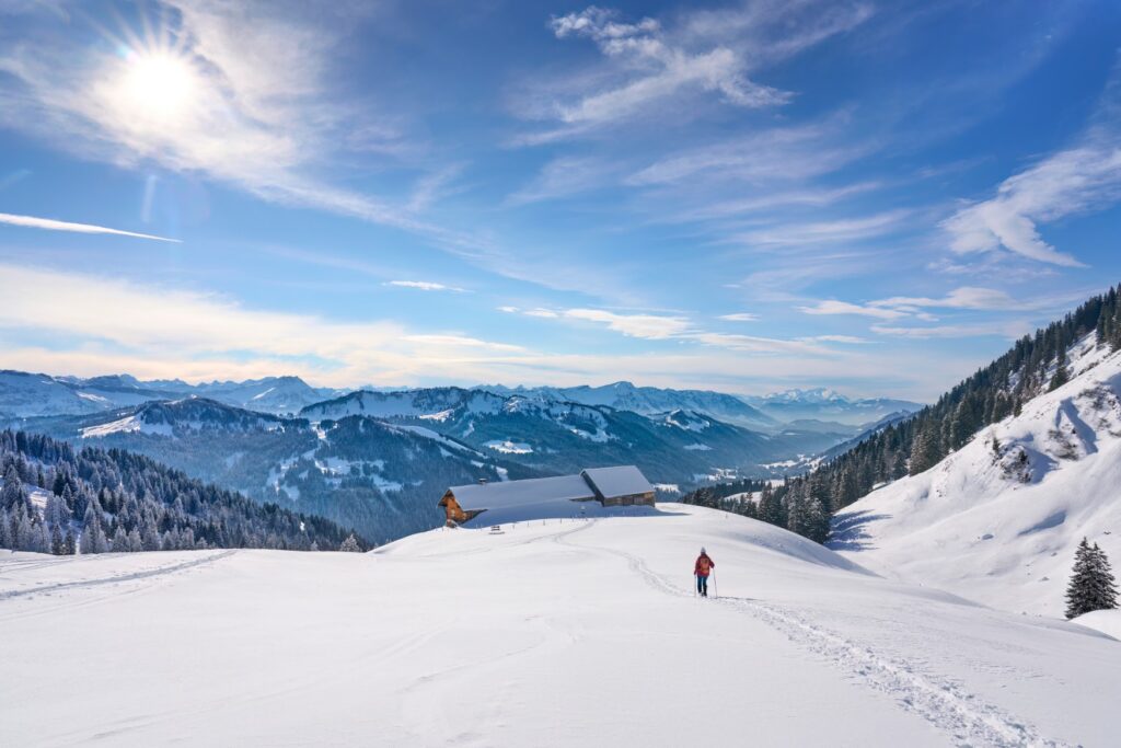 On a snow-covered slope in the mountains, a person dressed in winter clothing and carrying snowshoes runs downhill toward a hut on a ledge. Behind it, a mountain valley with lots of forest opens up, and the sky above is blue.