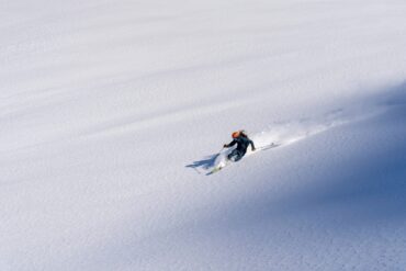 Drone footage of a skier speeding downhill through fresh, deep snow, leaving clouds of snow behind him.
