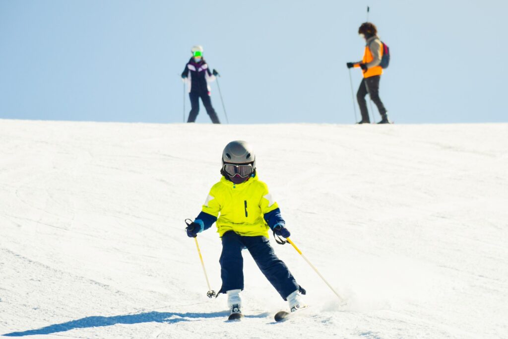 A child in neon yellow winter clothing is skiing down a slope directly toward the viewer. Two adults are standing at the top of the slope, with blue sky behind them.