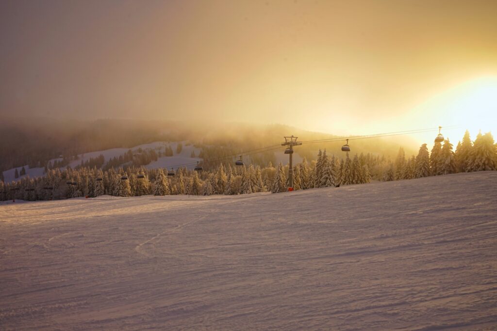 A ski chairlift stretches from left to right across a snow-covered slope. The mountain slope behind it is half covered by fog, through which the sun shines from the right.
