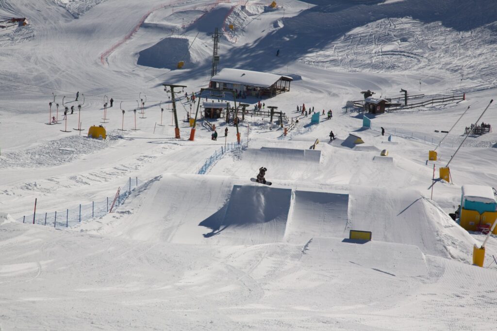 Drone footage of a snowboard course under thick snow. A snowboarder is flying over a ramp, with a chairlift and the corresponding station visible in the background.