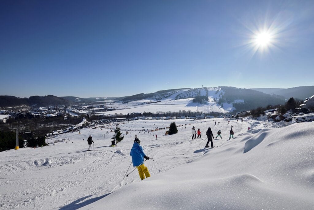 Über einen sanften, verschneiten Hang fahren dutzende Personen in Winterkleidung auf Skiern in ein Tal hinab. Links im Bild im Hintergrund eine Ortschaft, rechts erhebt sich ein bewaldeter Berg. Über allem strahlend blauer Himmel.