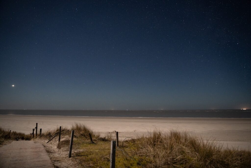 Vom Betrachter führt ein Steg aus Holzbohlen zu einem breiten, langen Sandstrand. Darüber liegt ein klarer Nachthimmel mit vielen sichtbaren Sternen.