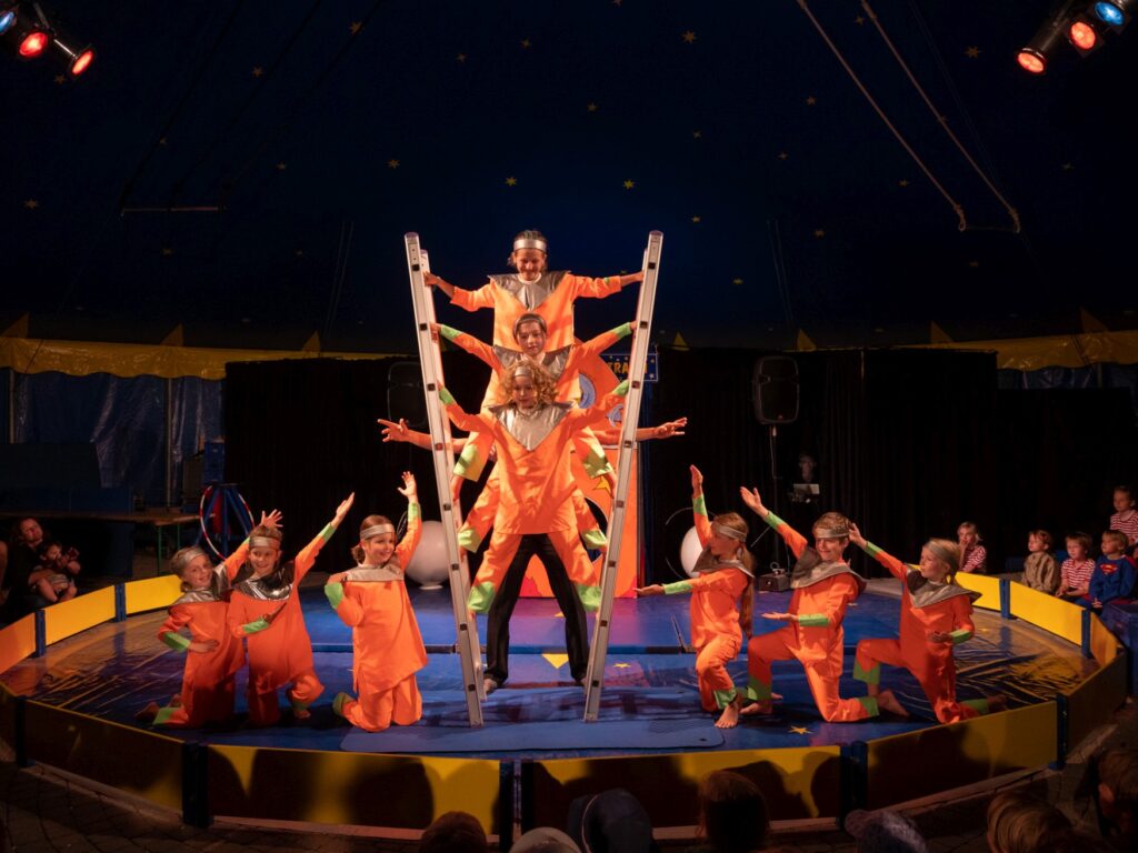 In einer kleinen Zirkusmanege balancieren drei Kinder in orangenen Anzügen zwischen zwei freistehenden Leiter. Links und rechts von ihnen hocken je drei weitere Kinder im gleichen Aufzug.