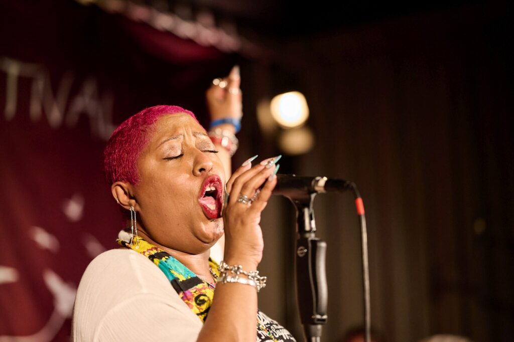 Eine Frau mit kurzen, rot gefärbten Haaren singt auf einer Bühne mit geschlossenen Augen in ein Mikrofon. Die andere Hand hat sie in die Höhe gestreckt.