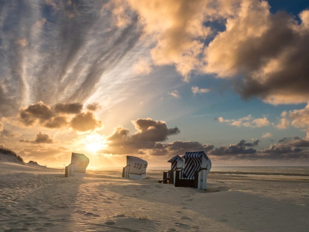 Auf einem breiten Strandabschnitt, rechts das Meer, stehen leicht versetzt fünf helle Strandkörbe im Sand. Im Hintergrund geht gerade die Sonne goldgelb zwischen einigen Wolken auf.