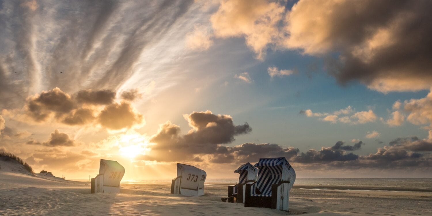 Auf einem breiten Strandabschnitt, rechts das Meer, stehen leicht versetzt fünf helle Strandkörbe im Sand. Im Hintergrund geht gerade die Sonne goldgelb zwischen einigen Wolken auf.