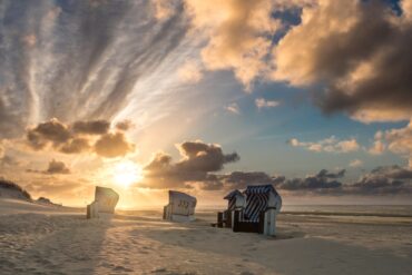Auf einem breiten Strandabschnitt, rechts das Meer, stehen leicht versetzt fünf helle Strandkörbe im Sand. Im Hintergrund geht gerade die Sonne goldgelb zwischen einigen Wolken auf.