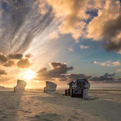 Auf einem breiten Strandabschnitt, rechts das Meer, stehen leicht versetzt fünf helle Strandkörbe im Sand. Im Hintergrund geht gerade die Sonne goldgelb zwischen einigen Wolken auf.