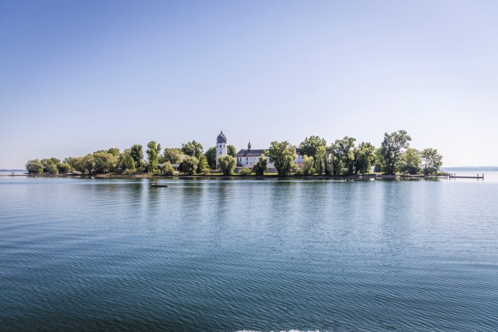 Eine grüne Insel liegt inmitten eines stillen Sees, darauf sichtbar ein weißes Kloster. Die Insel spiegelt sich in der Wasseroberfläche.