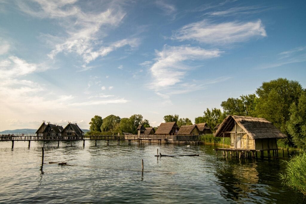 An einem grünen Seeufer stehen einige Nachbauten steinzeitlicher Pfahlbauten, größtenteils aus Holz und Reisig errichtet.