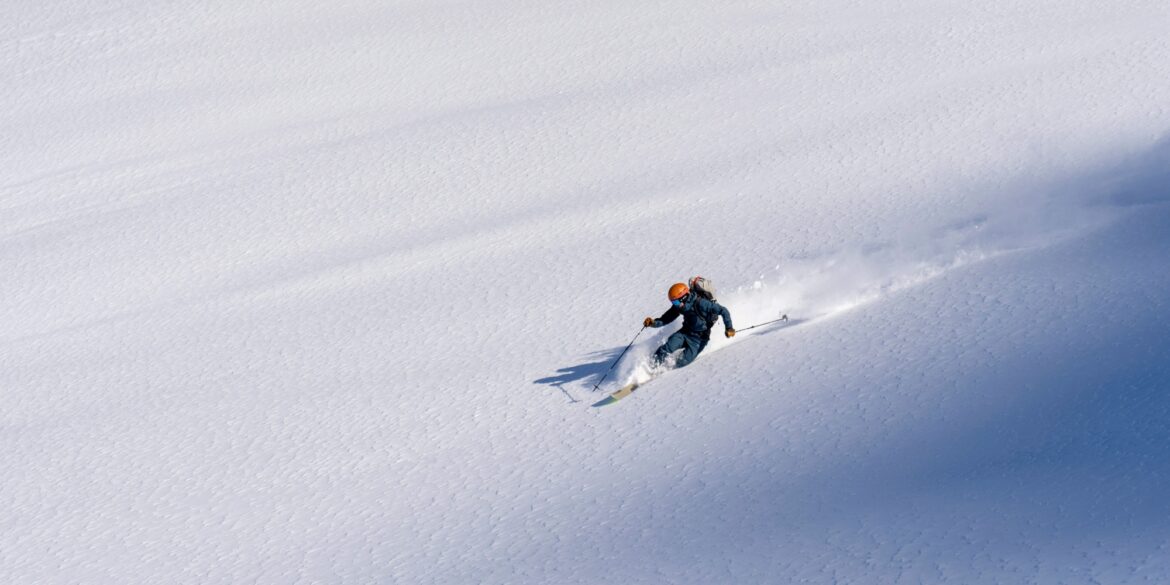 Drohnenaufnahme eines Skifahrers, der durch frischen, tiefen Schnee schnell hinabfährt und dabei Schneewolken hinter sich aufzieht.