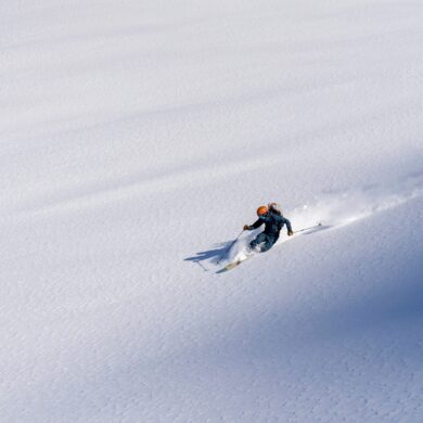 Drohnenaufnahme eines Skifahrers, der durch frischen, tiefen Schnee schnell hinabfährt und dabei Schneewolken hinter sich aufzieht.