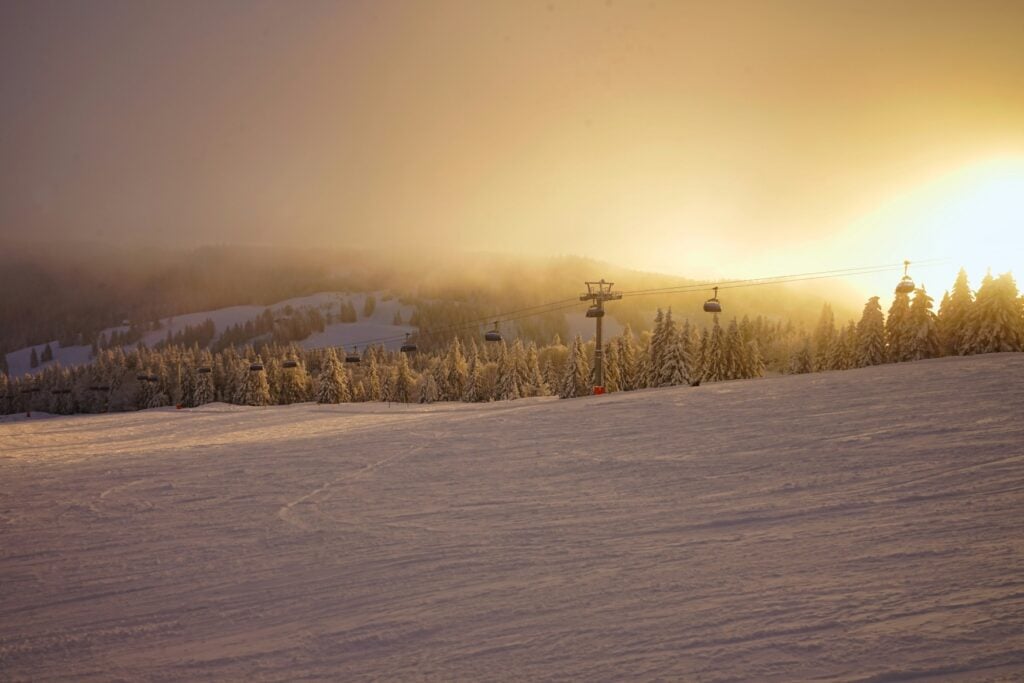 Über einen schneebedeckten Hang spannt von links nach rechts ein Ski-Sessellift. Der Berghang dahinter ist halb von Nebel verdeckt, durch den von rechts die Sonne strahlt.