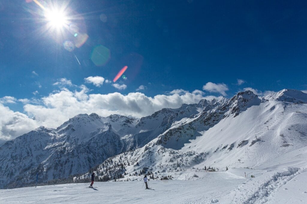 Zwei Personen in Wintersportkleidung fahren auf Skiern einen sachten Hang hinab in ein verschneites Tal. Im Hintergrund zieht eine Bergkette durchs Bild, darüber blauer Himmel.