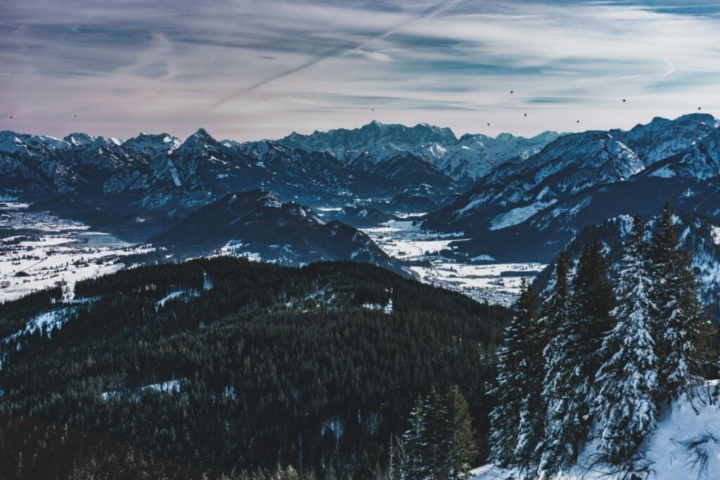 Panoramablick über die zerklüfteten Gipfel der alpen in einem kalten, blauen Winterlicht. Rechts am Horizont sind mehrere Heißluftballons erkennbar.
