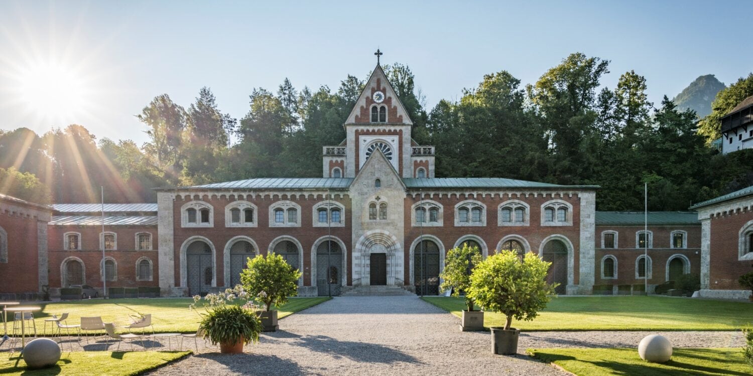 Ein neoromanischer Backsteinbau zieht sich quer durchs Bild, über dem mittigen Eingang geht ein kleine Turm nach oben. Vor dem Eingang erstreckt sich ein kleiner Park, der mittlere Weg führt genau auf das Gebäude zu. Darüber blauer Himmel und strahlender Sonnenschein.