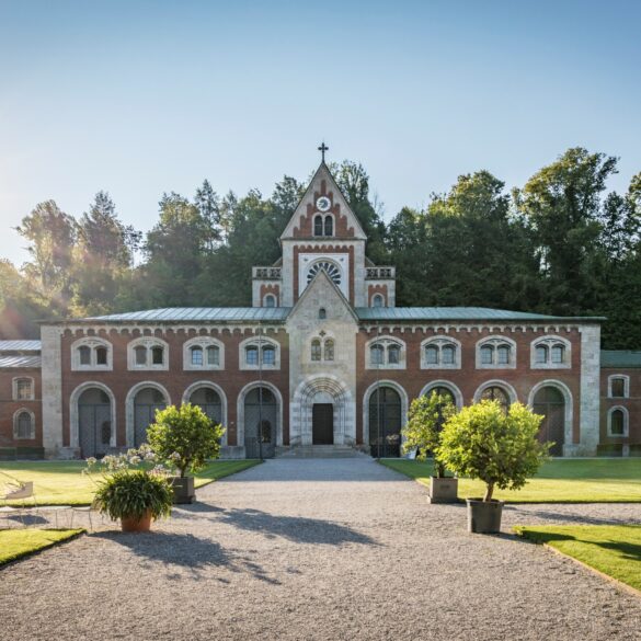 Ein neoromanischer Backsteinbau zieht sich quer durchs Bild, über dem mittigen Eingang geht ein kleine Turm nach oben. Vor dem Eingang erstreckt sich ein kleiner Park, der mittlere Weg führt genau auf das Gebäude zu. Darüber blauer Himmel und strahlender Sonnenschein.