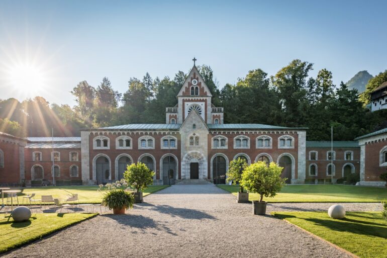 Ein neoromanischer Backsteinbau zieht sich quer durchs Bild, über dem mittigen Eingang geht ein kleine Turm nach oben. Vor dem Eingang erstreckt sich ein kleiner Park, der mittlere Weg führt genau auf das Gebäude zu. Darüber blauer Himmel und strahlender Sonnenschein.