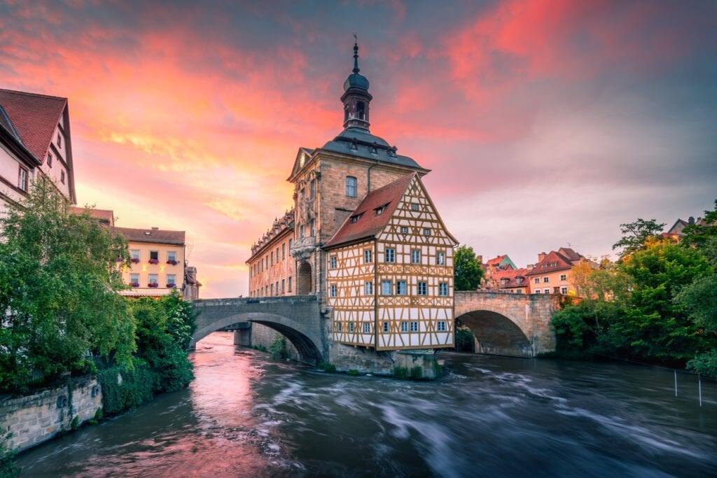 Quer über einen Fluss spannt eine steinerne Brücke, darauf ein hohes Barockgebäude mit Fachwerkfassade.