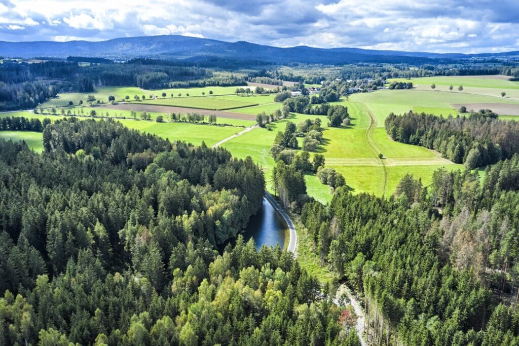 Drohnenaufnahme eines grünen Flusstals mit mehreren Waldstücken, rechts im Hintergrund eine kleine Siedlung und ganz im Hintergrund ein bewaldeter Höhenzug, darüber dichte Wolken.