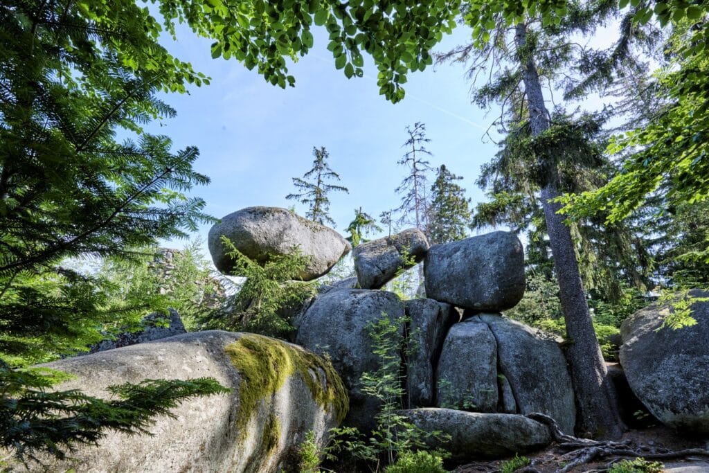 In einem grünen Wald liegen mehrere große Felsbrocken übereinander, darüber blauer Himmel.