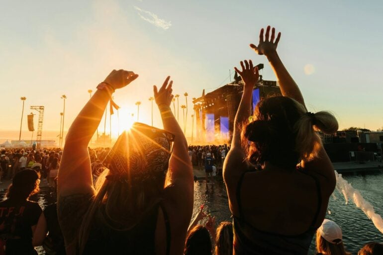 Two women in summer clothes stand with their backs to the camera and stretch their arms toward the sky. In front of them stands a large crowd in front of a festival stage, above which the evening sky is gently illuminated by the sunset.
