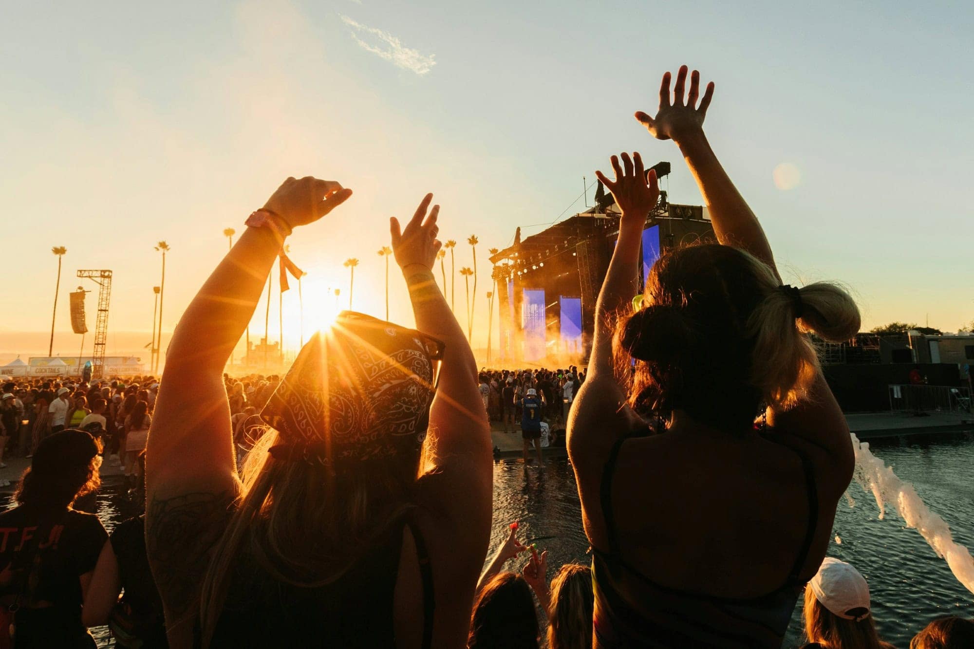 Two women in summer clothes stand with their backs to the camera and stretch their arms toward the sky. In front of them stands a large crowd in front of a festival stage, above which the evening sky is gently illuminated by the sunset.