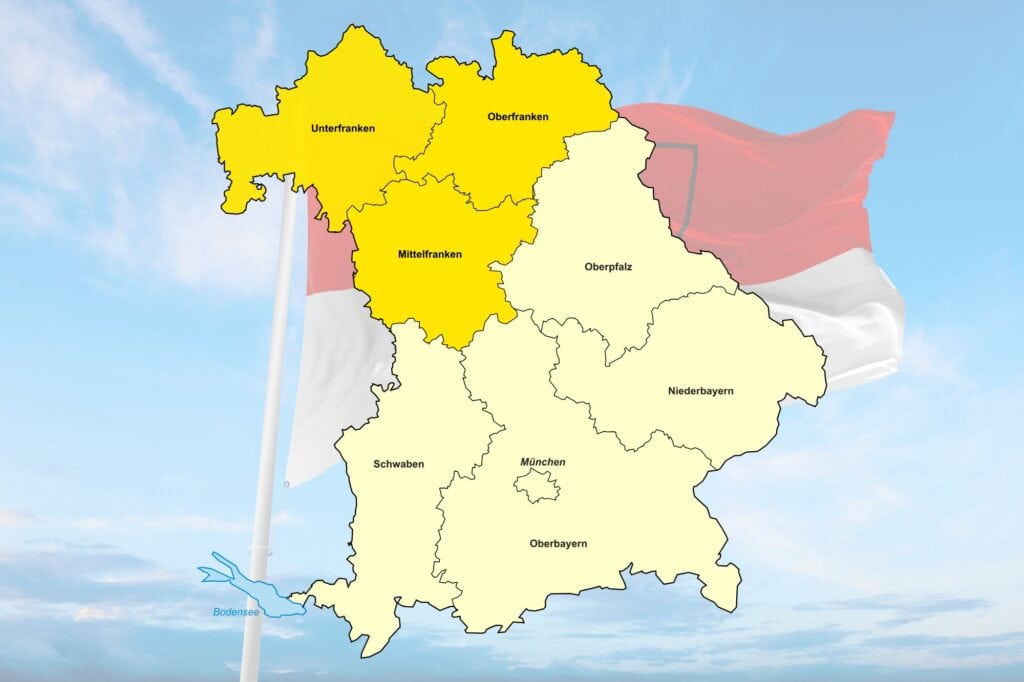 Eine freigestellte Karte des Bundeslands Bayern zeigt hervorgehoben die drei Regierungsbezirke Frankens, dahinter die rot-weiße Flagge Frankens vor blauem Himmel.