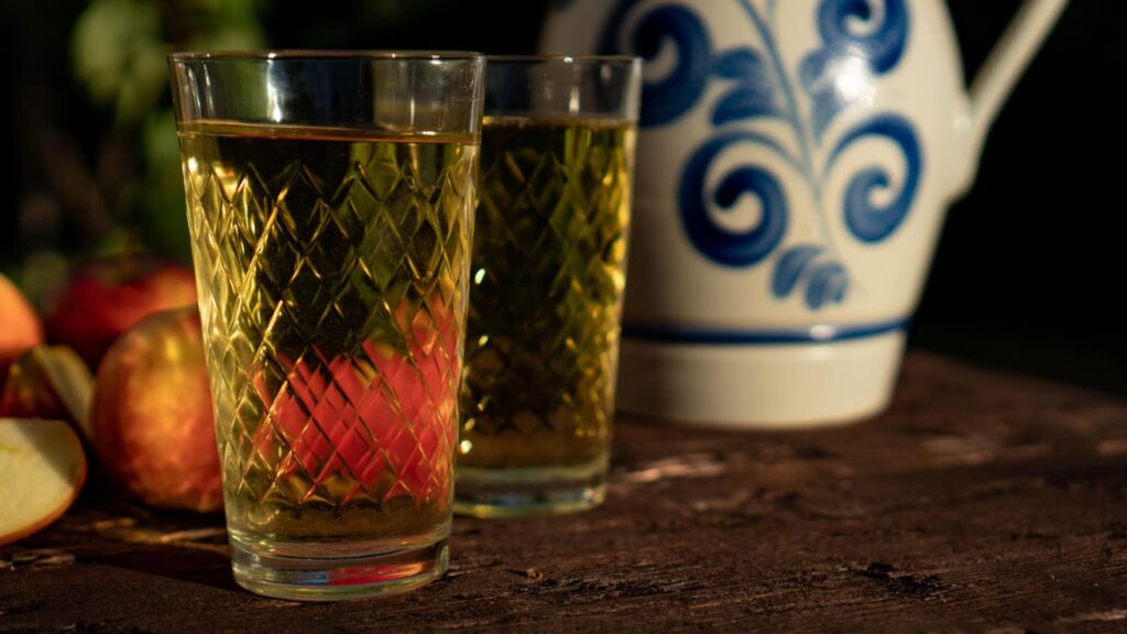 Close-up of two “ribbed” glasses of apple wine on a wooden table, with some apples and a clay “Bembel” jug slightly out of focus in the background.