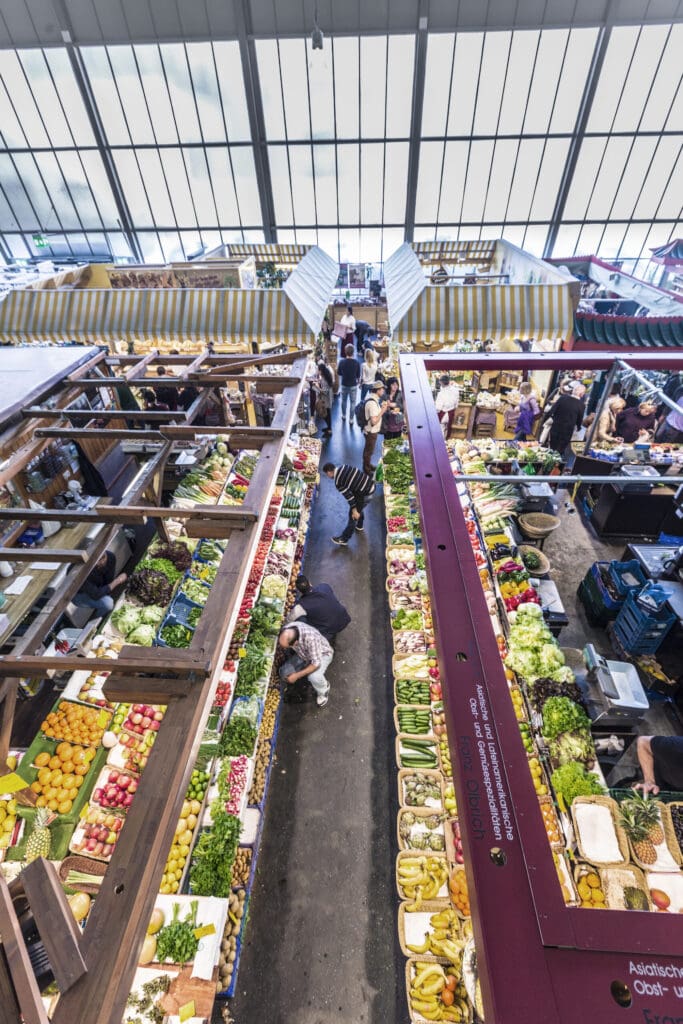 In a hall with a glass roof, numerous market stalls stand close together, with lots of fruit and vegetables on sale. A few people move through the aisle between the two rows of stalls.