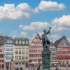 A marketplace with a row of colorful half-timbered houses. There are many people walking around the square. The focus is on a tall statue of Lady Justice.