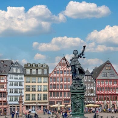A marketplace with a row of colorful half-timbered houses. There are many people walking around the square. The focus is on a tall statue of Lady Justice.