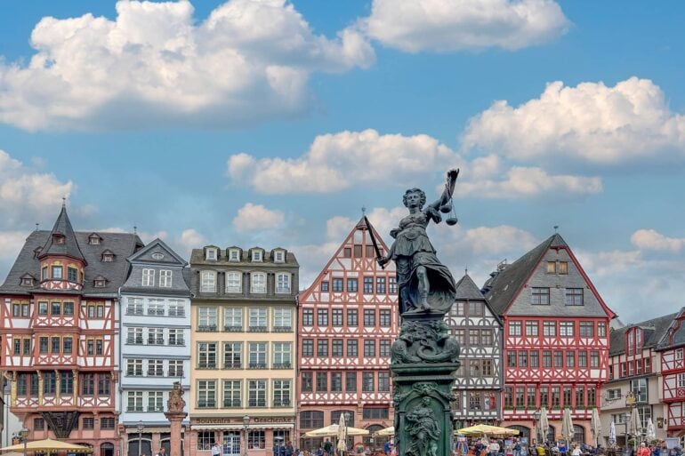 A marketplace with a row of colorful half-timbered houses. There are many people walking around the square. The focus is on a tall statue of Lady Justice.