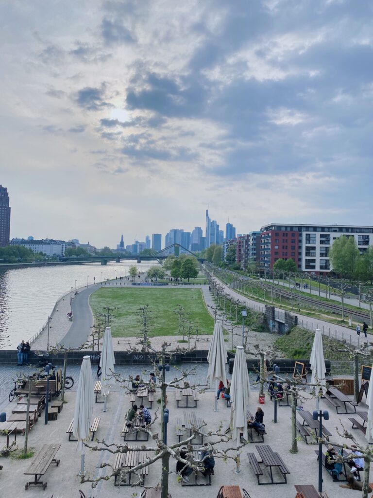 A concrete area with many seats, photographed from a high vantage point. There are many people on the area. In the background, there is a meadow and, on the left, a river stretching to the horizon. There, behind a light blue haze, the Frankfurt skyline can be seen.