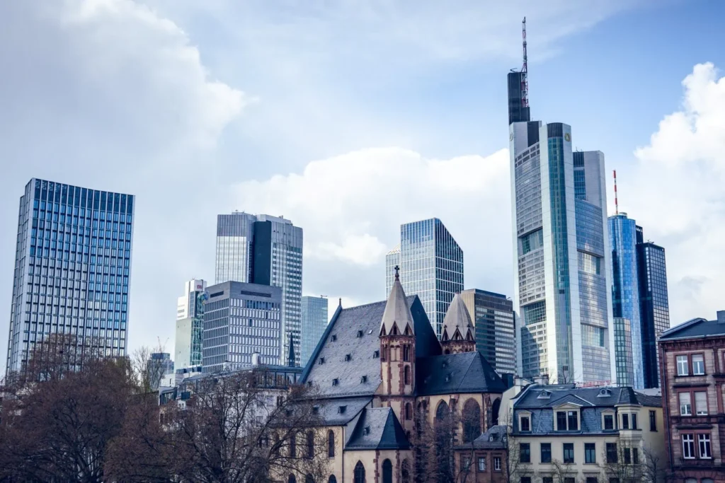 A small church made of light sandstone with a black roof stands in front of five high-rise buildings on the Frankfurt skyline.
