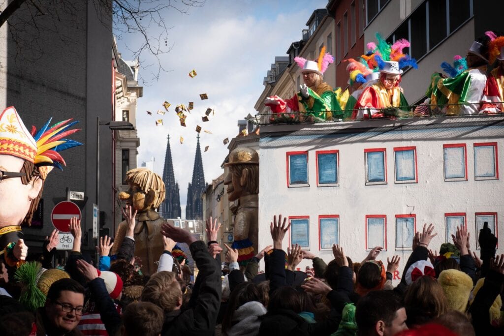 Several costumed people on a carriage thats disguised as a paper house throw sweets into a crowd of people in front of them. In the background between two houses appear the two towers of the Cologne Cathedral.