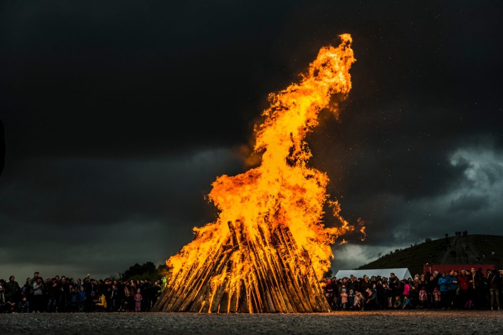A huge fire from a wooden pile burns bright in a dark night. Behind it is a huge crowd of onlookers.