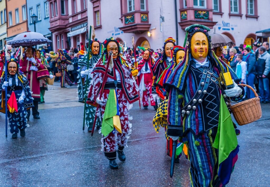 Several people in colorful costumes and with traditional carnival masks walk across a street.